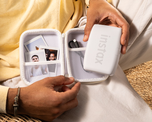Mani tengono una custodia bianca aperta con foto, caricabatterie e una stampante Instax bianca.