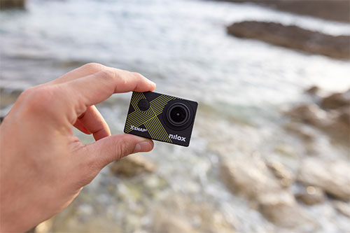 Una mano tiene una action camera nera con dettagli gialli. Sullo sfondo, acqua e rocce.