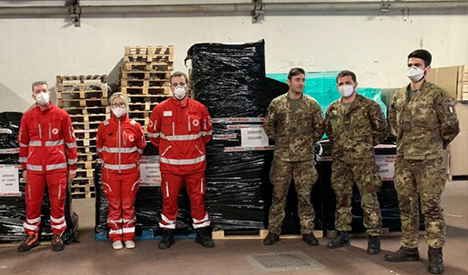 Gruppo di sei persone in uniforme rossa e mimetica con mascherine, davanti a bancali e pacchi avvolti in plastica nera.