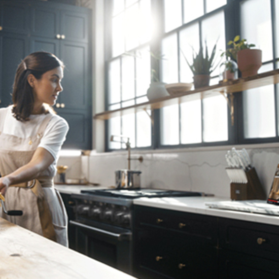 Donna in cucina con grembiule chiaro, mobili blu scuro, grande finestra a riquadri e ripiano con vasi di piante.