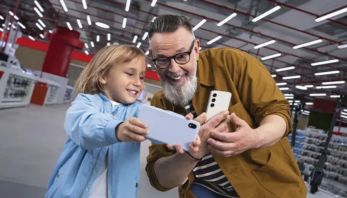 Un uomo e una bambina sorridono tenendo in mano dei telefoni in un negozio con scaffali e luci.