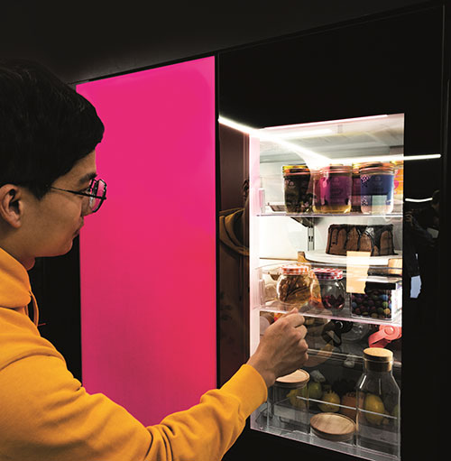 Un uomo in felpa gialla apre un frigorifero nero con ripiani illuminati pieni di barattoli e bottiglie. La porta � rosa.