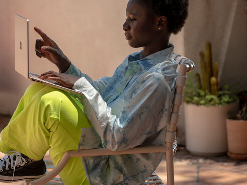 Donna seduta su sedia usa un laptop, pantaloni verdi, camicia azzurra. Piante in vasi sullo sfondo.