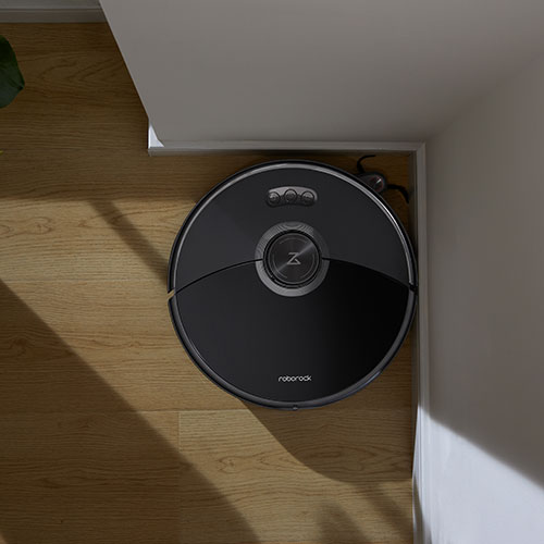 Aspirapolvere robot nero su pavimento in legno chiaro, vicino a parete bianca.