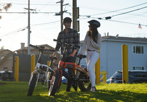 Uomo e donna in piedi con bici su erba. L'uomo indossa una camicia a quadri e la donna indossa un casco.