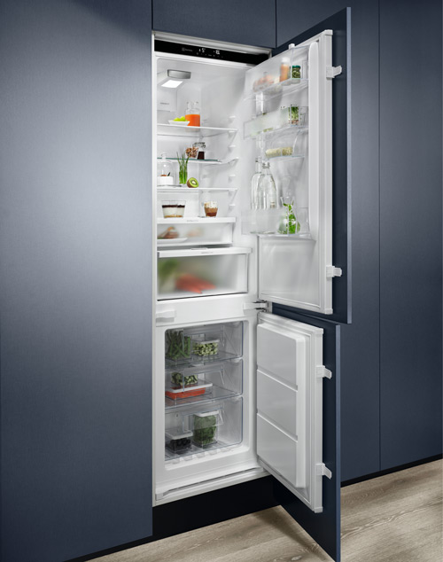 Frigorifero bianco aperto, integrato in mobili blu. Ripiani interni con cibo e bevande visibili. Pavimento chiaro.