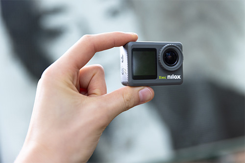 Primo piano di una mano che tiene una action camera grigia con schermo e obiettivo visibili. Sul corpo macchina è presente la scritta "nilox". Lo sfondo è sfocato.