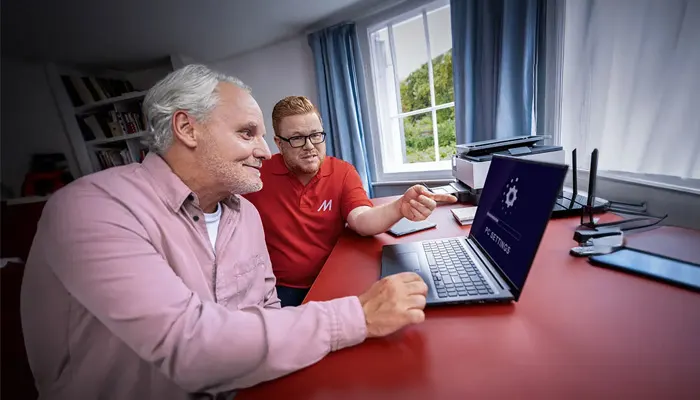 Uomini davanti a un portatile su una scrivania rossa. Un uomo indica lo schermo. Sullo sfondo, finestra, libreria, stampante e router.