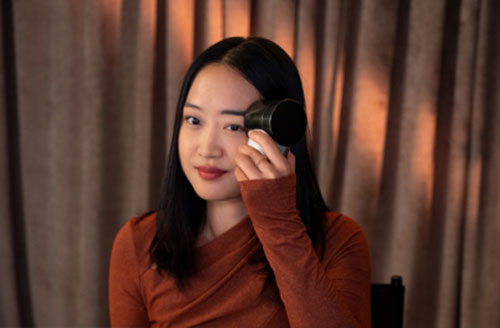 Donna con maglia arancione applica trucco. Sfondo con tende marroni.