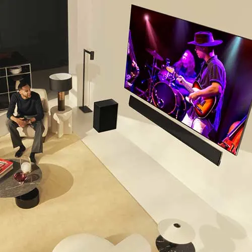 Uomo seduto su una poltrona guarda la TV che mostra una band musicale. Altri mobili e altoparlanti visibili nella stanza.