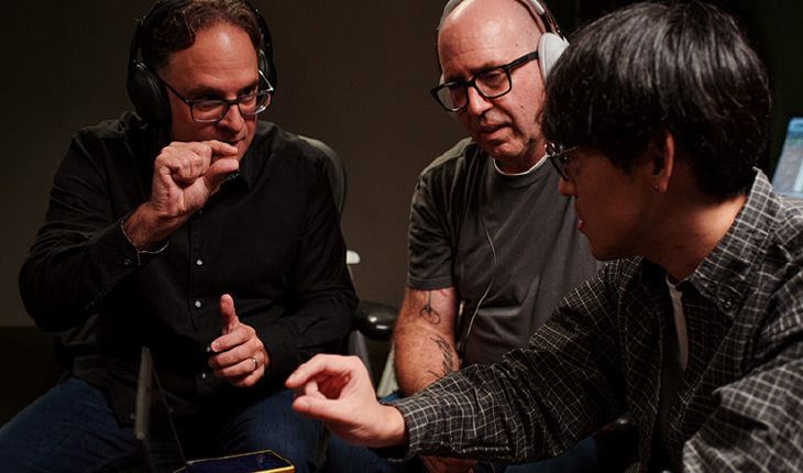 Tre professionisti dell'audio in studio, con cuffie e occhiali, concentrati in una discussione tecnica mentre analizzano dei dettagli gesticolando con le mani.