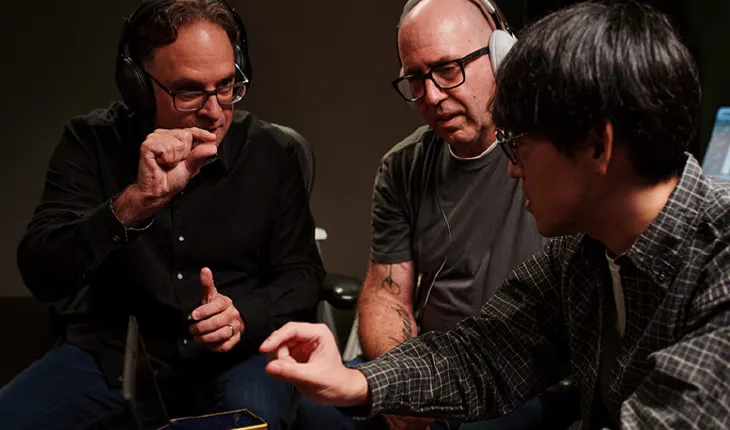 Tre professionisti dell'audio in studio, con cuffie e occhiali, concentrati in una discussione tecnica mentre analizzano dei dettagli gesticolando con le mani.