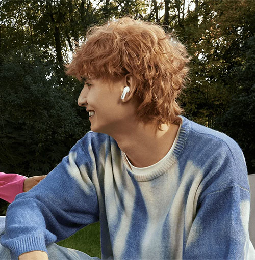 Uomo sorridente con auricolare bianco, indossa maglione blu e bianco. Sfondo con alberi verdi.