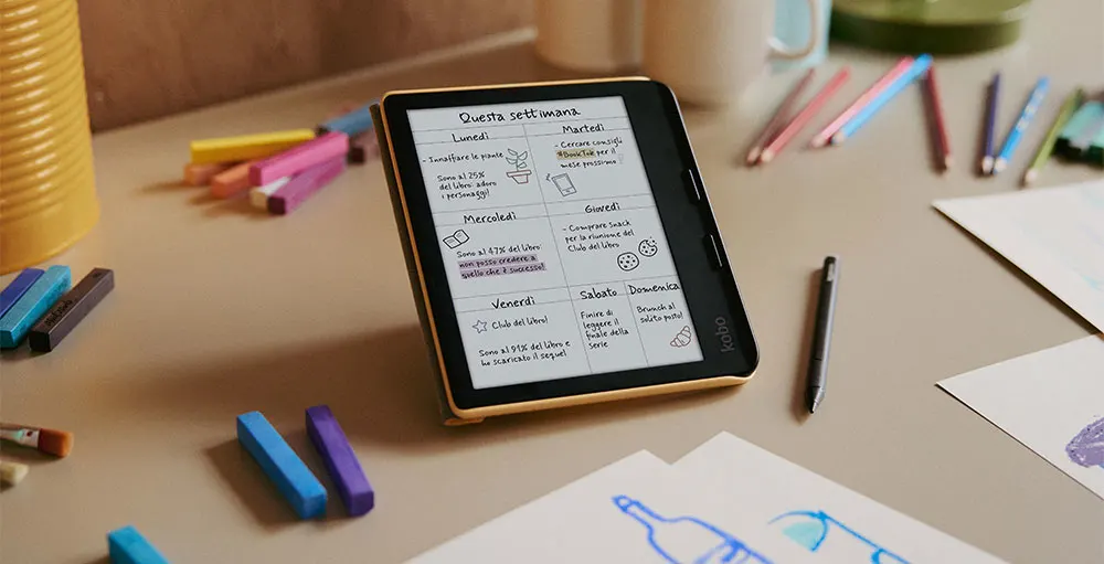 E-reader con agenda settimanale, pennarello e pastelli colorati su tavolo. Fogli con disegni sparsi.