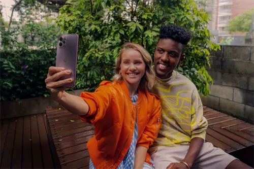 Due persone sorridono mentre una scatta un selfie con un telefono viola su una panchina di legno, con alberi e un edificio sullo sfondo.