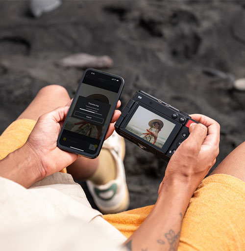 Mani tengono un telefono e una fotocamera su una spiaggia scura. Il telefono mostra una barra di caricamento. La fotocamera mostra una foto.
