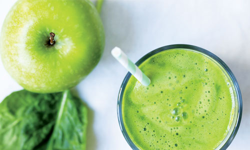 Su sfondo chiaro, un bicchiere di vetro contiene un frullato verde con cannuccia, accanto a una mela verde e foglie di spinaci.