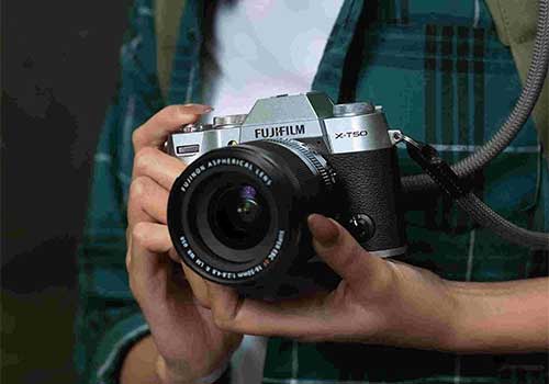 Una persona tiene in mano una fotocamera Fujifilm X-T50 argento e nera con un obiettivo grande. Ha una tracolla grigia.