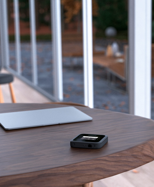 Su tavolo di legno, un laptop grigio e un dispositivo nero quadrato. Dietro, ringhiera bianca e vegetazione sfocata.