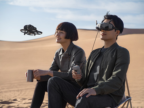 Uomo con visore VR e donna con tazza in deserto, seduti. Un drone nero vola in alto. Cielo azzurro sullo sfondo.