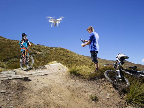 Ciclisti in montagna con drone. Uno controlla il drone, l'altro in bici. Bicicletta a terra con casco. Cielo azzurro.