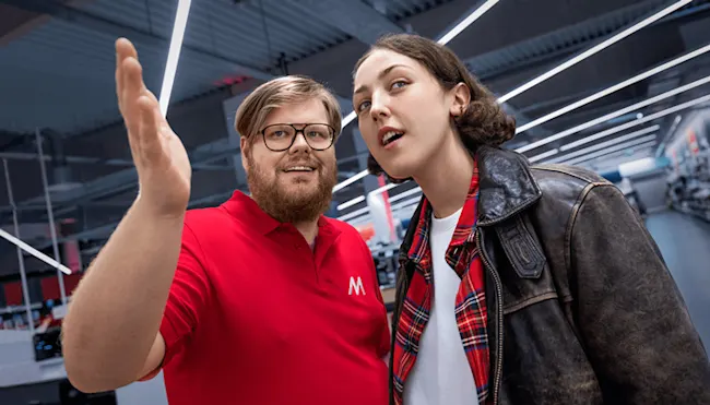 Uomo in maglia rossa saluta, donna in giacca di pelle accanto. Sullo sfondo, luci lineari e scaffali di un negozio.