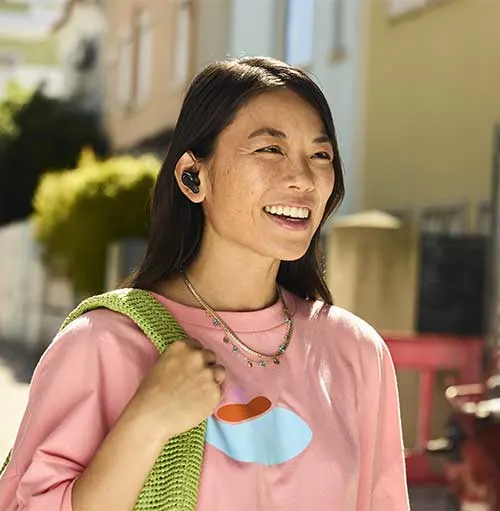 Donna sorridente con auricolari neri, indossa maglia rosa, collana e borsa verde. Sfondo urbano sfocato.