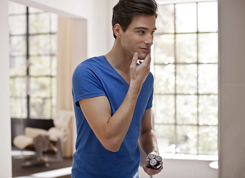 Uomo in t-shirt blu tocca il mento, tiene un rasoio elettrico. Finestra con griglia sullo sfondo, stanza con mobilio.