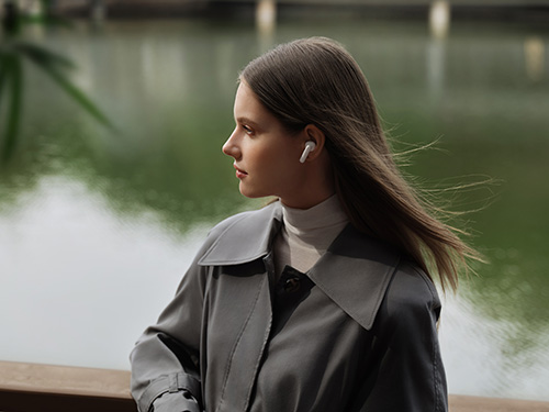 Donna con cappotto grigio e auricolare, rivolta verso sinistra. Sfondo sfocato con acqua verde e ponti bianchi.