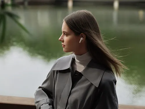 Donna con cappotto grigio e auricolare, rivolta verso sinistra. Sfondo sfocato con acqua verde e ponti bianchi.