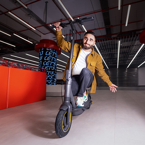 Uomo su monopattino grigio con accenti gialli. Indossa giacca ocra, pantaloni grigi e scarpe da ginnastica. Sfondo industriale con luci al neon.