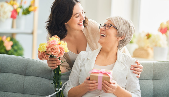 Madre sorride con regalo e fiori, abbracciata dalla figlia su divano grigio. Fiori e scaffale visibili sullo sfondo.