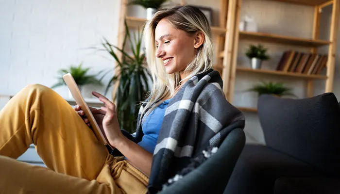Donna seduta su una sedia con una coperta, che utilizza un tablet. Sullo sfondo, una libreria in legno e piante.