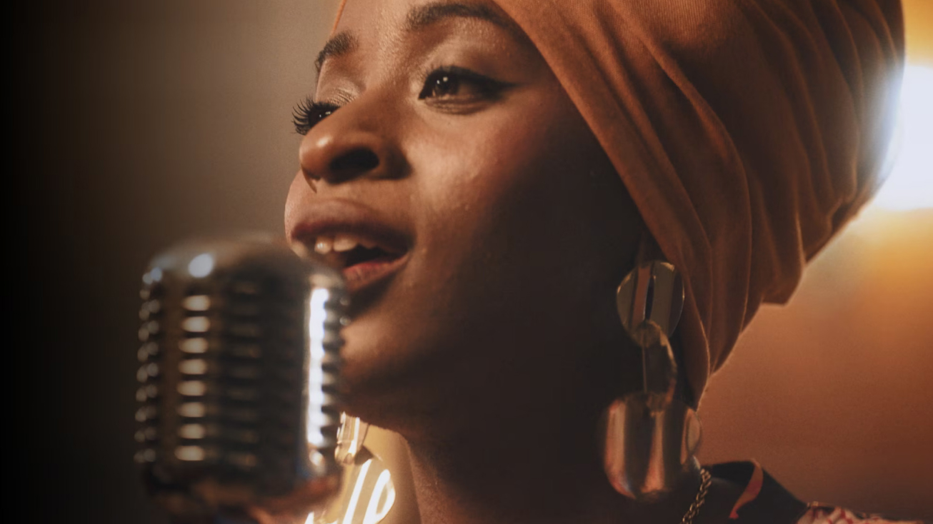 Cantante con turbante arancione vicino a microfono vintage. Indossa orecchini pendenti. Sfondo scuro con luci calde.