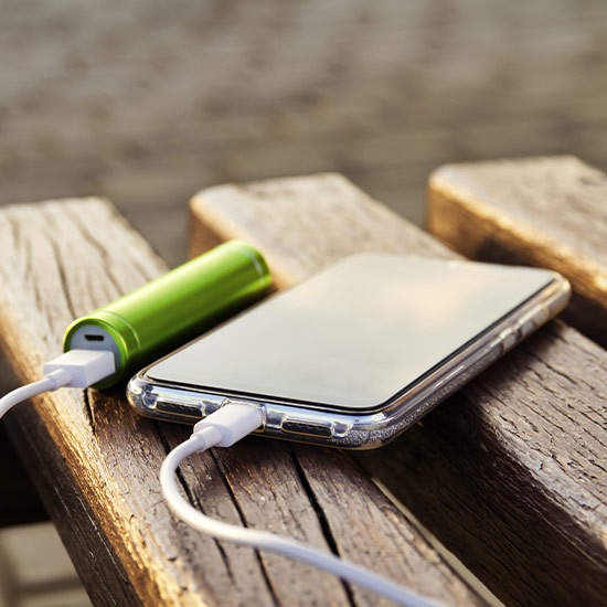 Su una panca di legno c'è un telefono collegato a una batteria esterna verde tramite un cavo bianco.