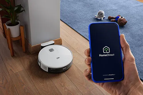 Aspirapolvere robot bianco su legno, caricatore, pianta, telefono con app HomeDirect e peluche su tappeto blu.