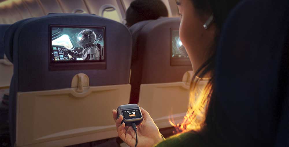 Interno aereo: passeggero tiene custodia auricolari, schermi mostrano astronauta. Sedili blu e illuminazione interna.