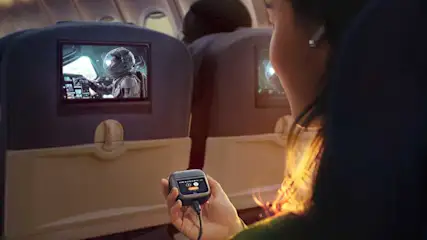 Interno aereo: passeggero tiene custodia auricolari, schermi mostrano astronauta. Sedili blu e illuminazione interna.