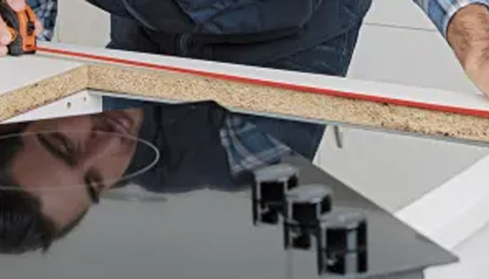 Un uomo misura un piano cottura nero su un ripiano di legno con un metro arancione. Il piano cottura riflette il suo viso.