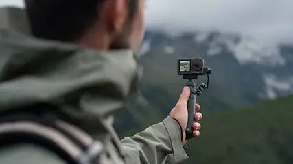 Uomo con zaino riprende montagne con action camera nera su impugnatura. Display acceso. Sfondo nebbioso.