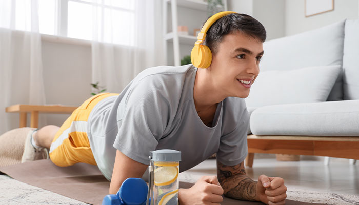 Un uomo con cuffie gialle fa esercizi plank su un tappeto. Accanto ci sono pesi e una bottiglia d'acqua. Dietro c'è un divano.