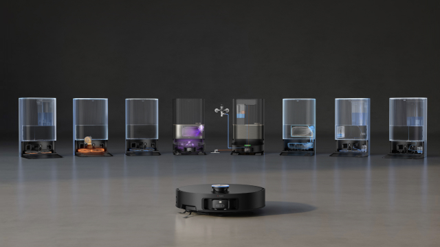 Un aspirapolvere robot nero di fronte a sette contenitori trasparenti su una superficie grigia. Lo sfondo è nero.