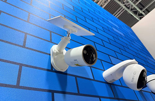 Telecamere bianche su muro di mattoni blu, una con pannello solare. Struttura metallica visibile sullo sfondo.