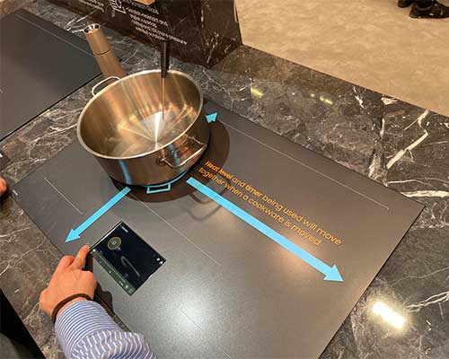 Pentola d'acciaio su piano cottura nero con frecce blu. Una mano indica un display nero.