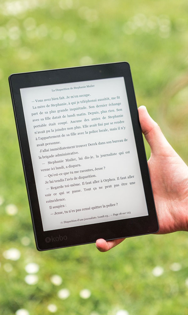 Una mano tiene un e-reader Kobo nero che mostra testo francese su sfondo verde sfocato.