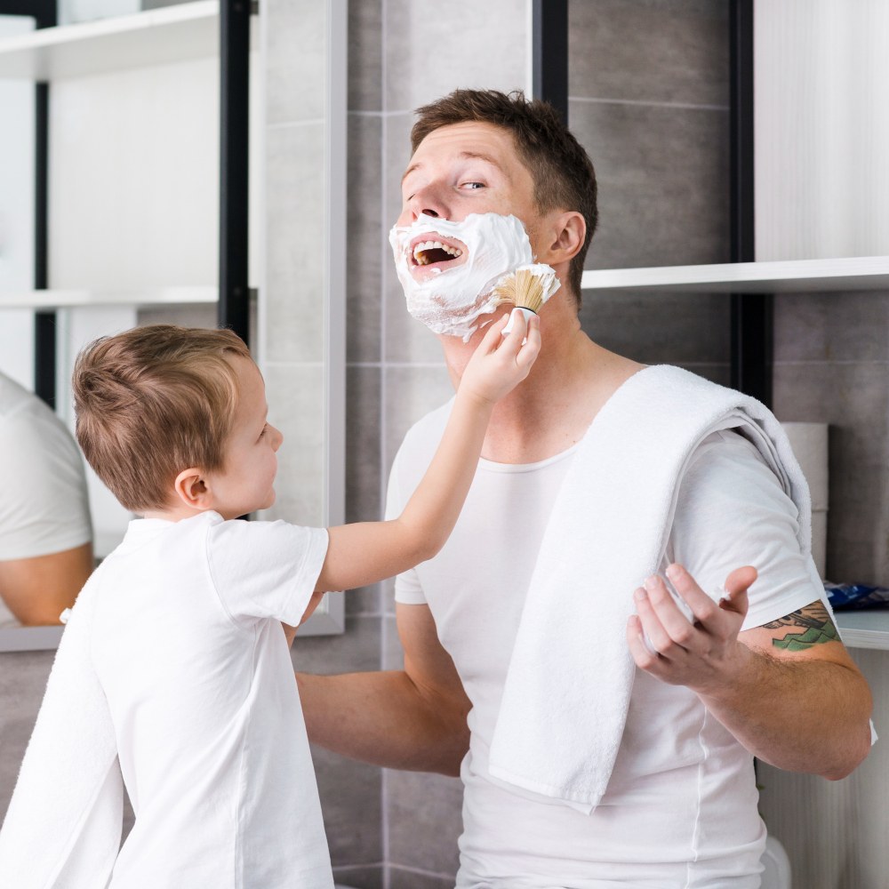 Padre con schiuma da barba sul viso. Bambino applica altra schiuma con pennello. Asciugamano bianco sulle spalle del padre.