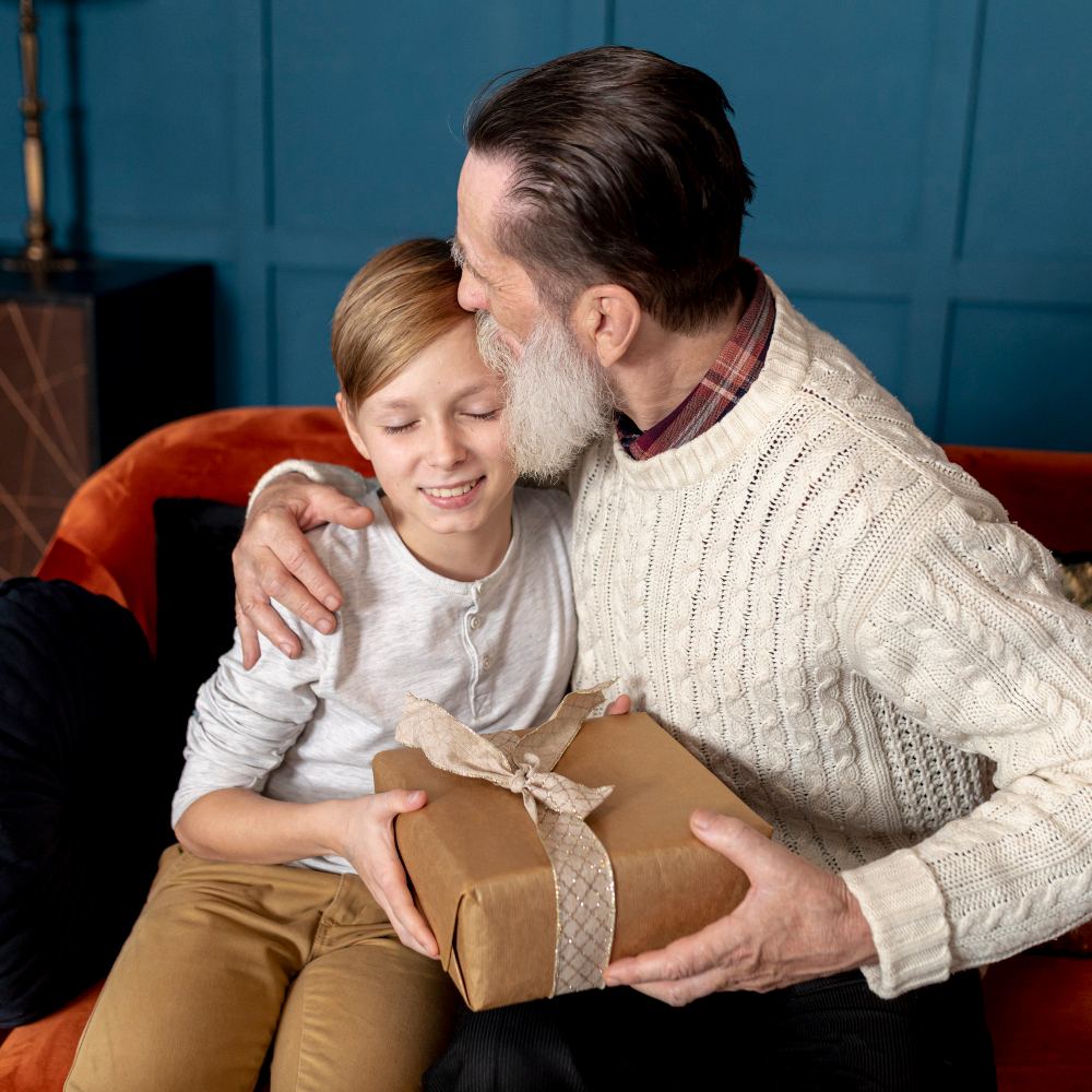 Un nonno abbraccia un bambino su un divano arancione mentre gli porge un regalo incartato con un fiocco.