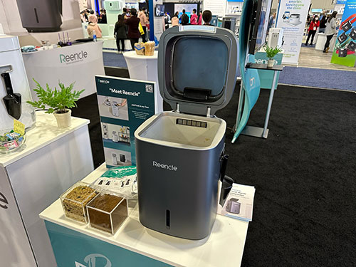 Un contenitore grigio con coperchio aperto su un tavolo bianco, accanto a piante e materiale informativo in un'area espositiva.