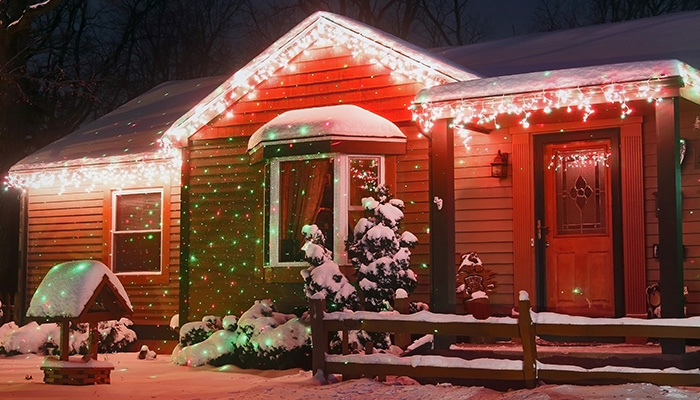 Casa in legno decorata con luci bianche e verdi, coperta di neve. Porta rossa con decorazioni natalizie.