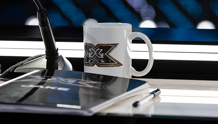 Foto della scrivania di Xfcator, con una tazza con logo Xfactor, un microfono e un copione.